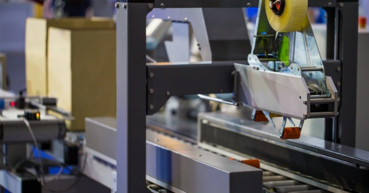 A brown cardboard box sits on the metal conveyor system of a carton sealing machine inside a warehouse.