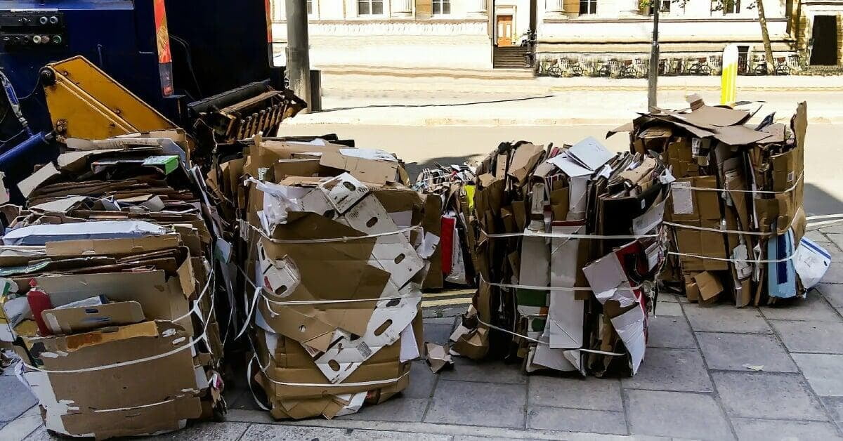 A row of bundled cardboard stacks beside a blue baling machine on a paved sidewalk near a city street.