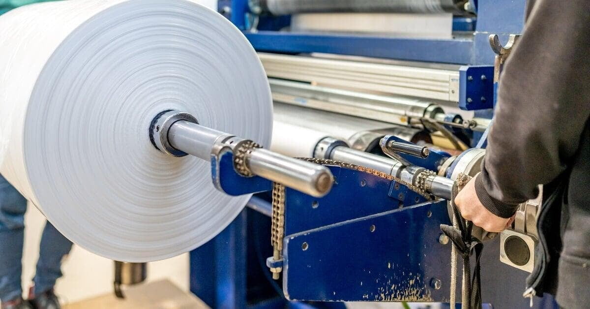 A large roll of white material mounted on blue industrial machinery with a person standing beside it.