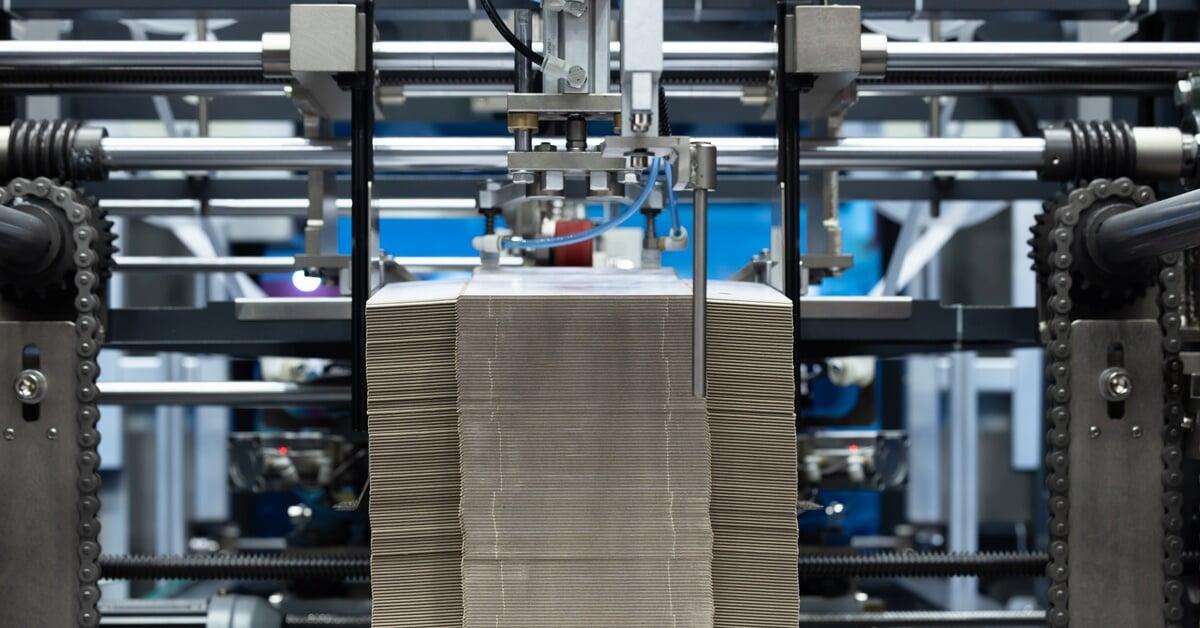 A stack of flattened pieces of material moving under a machine head inside an industrial production line.