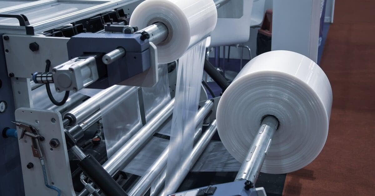 Two large white plastic film rolls feeding clear material through metal rollers on industrial machinery.