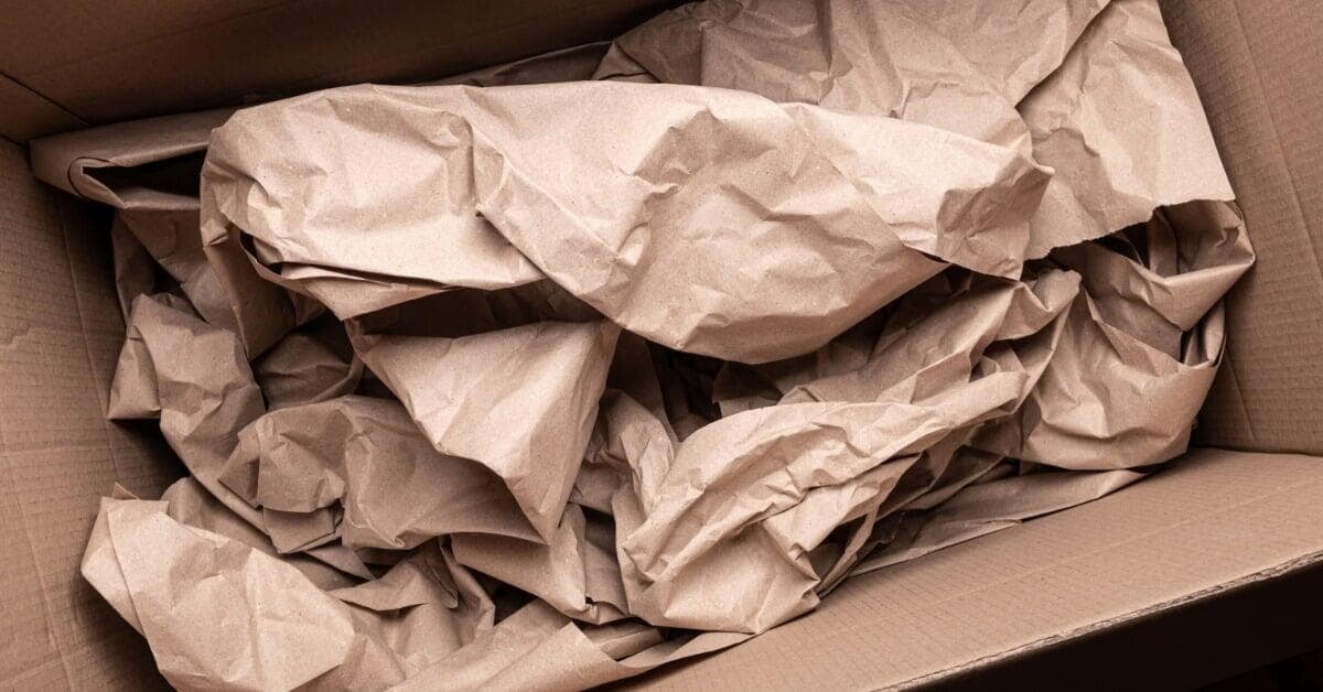 An open cardboard box filled with crumpled brown paper, with folded flaps around the packing material.