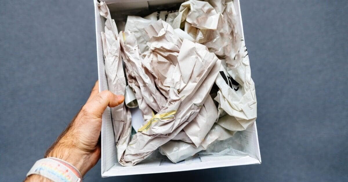 A hand holding a white box filled with crumpled newspaper and paper packing against a gray background.