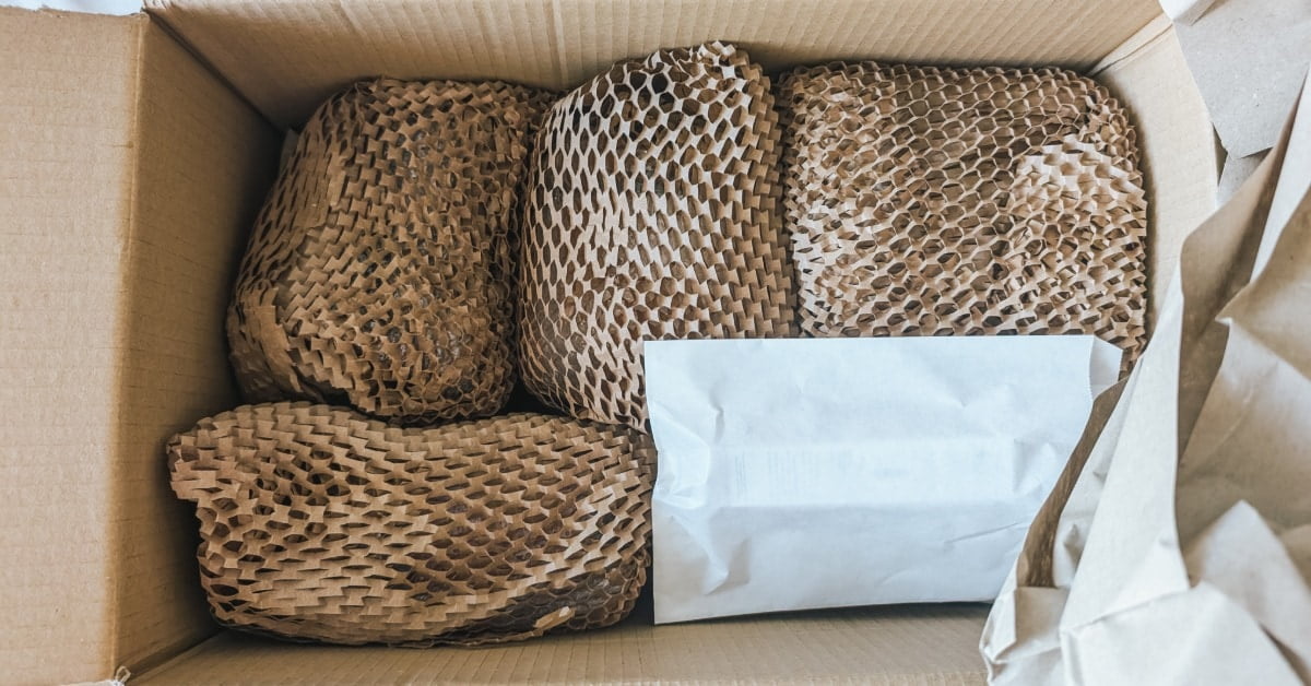 An open cardboard box holding wrapped items in honeycomb paper, white packaging, and crumpled paper sheets.