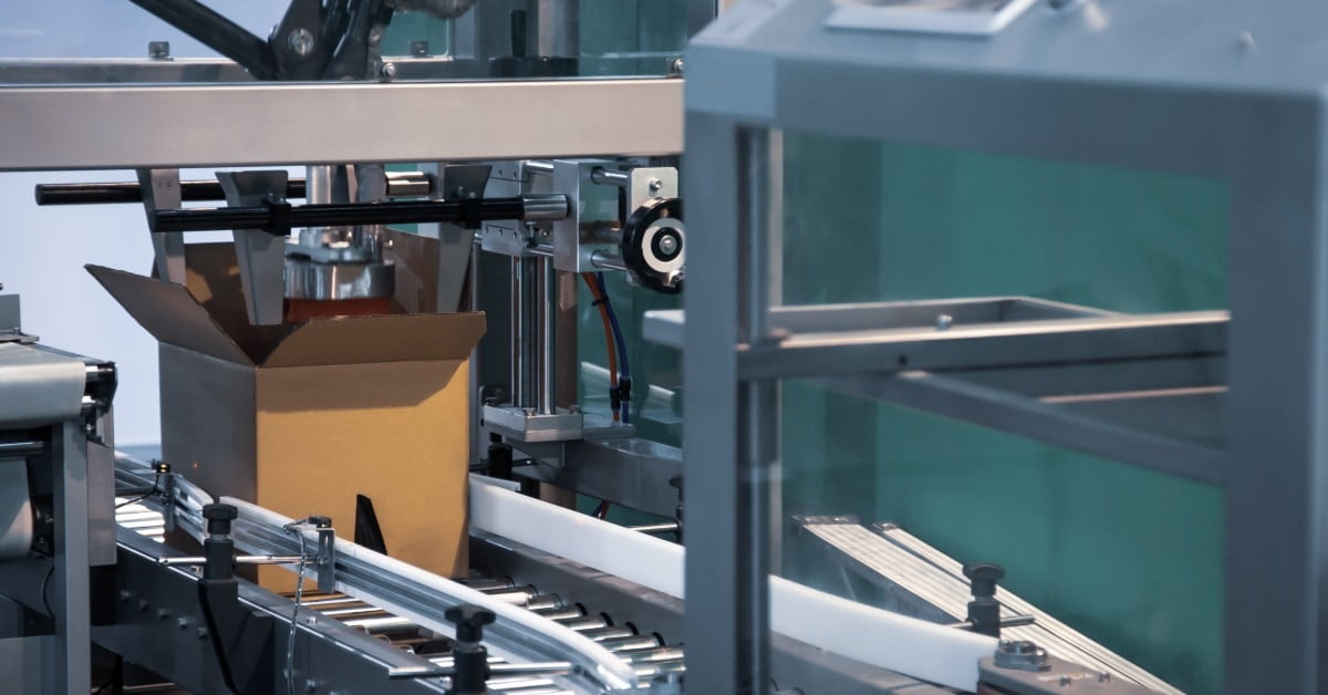 An open cardboard box moving through automated forming equipment with metal rails, guards, and control panels.