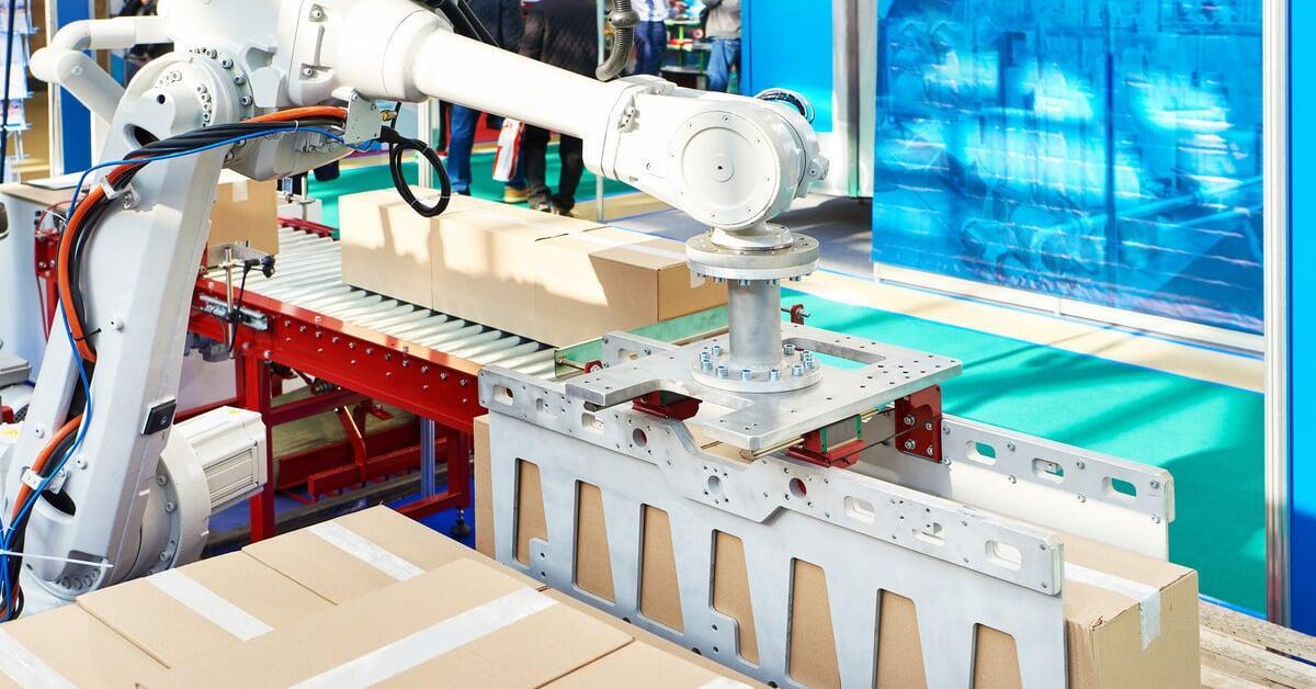 A robotic arm holding a package over a wooden pallet. More boxes are on another pallet and on the conveyor beside the robot arm.