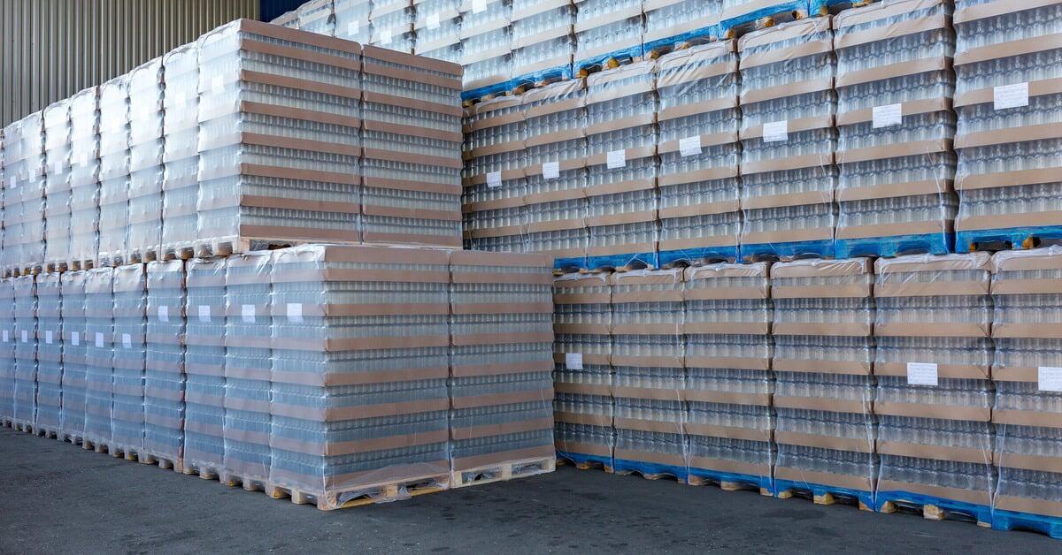 Dozens of wrapped pallets inside a warehouse. The front row is stacked two pallets high, and the back row is three pallets high.