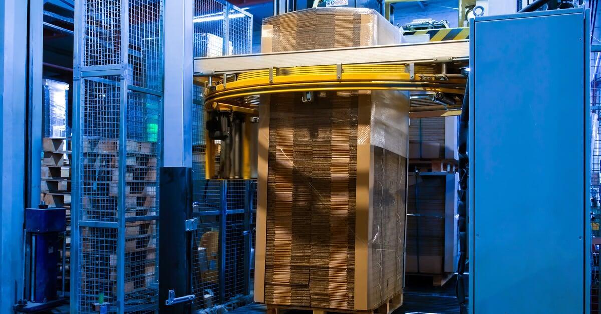 A yellow mechanical stretch wrapping ring encircling a tall stack of palletized cardboard. The stack is partially wrapped.