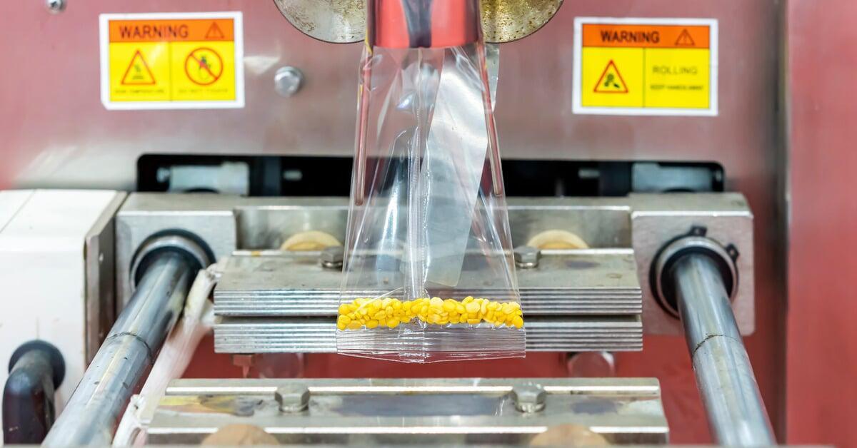 A bagging machine filling an open plastic bag with dry, yellow granules. Two warning stickers are on the machine.