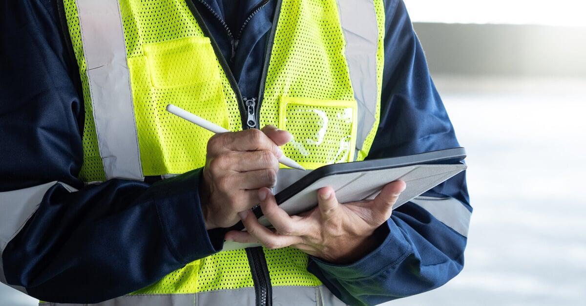 The midsection of a worker wearing a yellow hi-vis vest. They write a note on a tablet with a stylus.