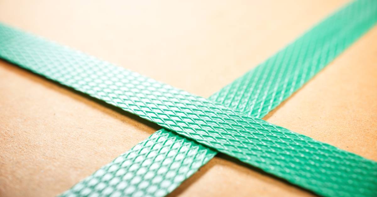 A close-up of two green embossed polyester straps. The straps crisscross on top of brown cardboard packaging.