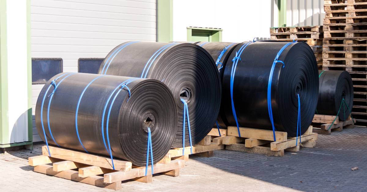 Large rolls of black industrial plastic strapped to wooden pallets. Some rolls are in the shade and some are in sunlight.