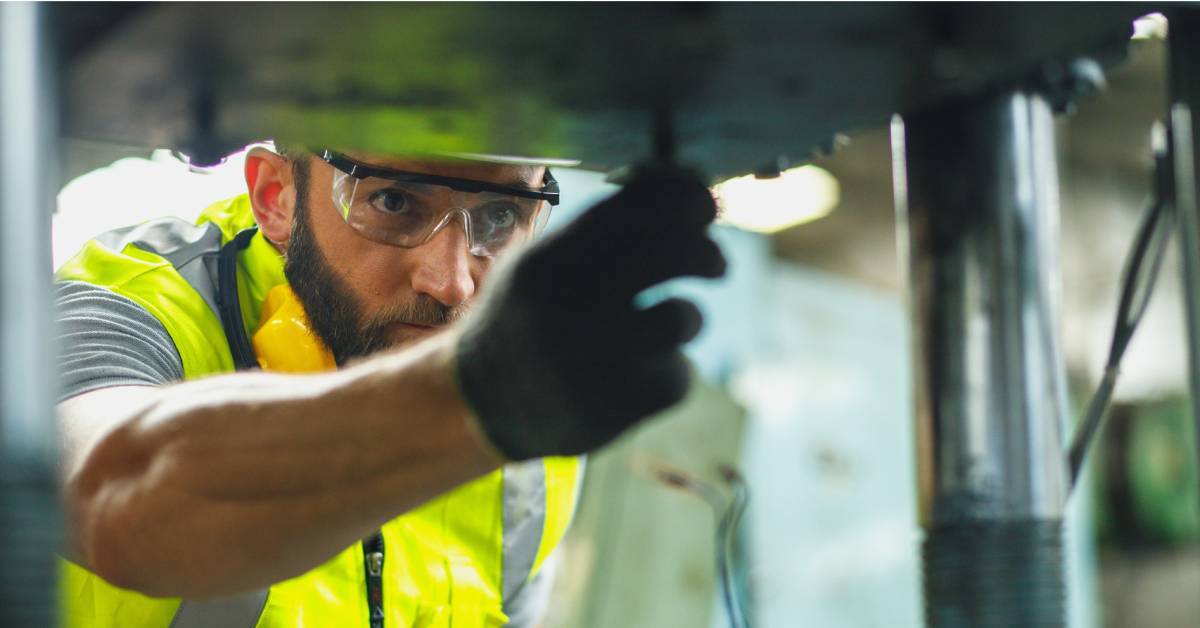 A worker pressing a tool against the underside of a metal surface. They wear a yellow hi-vis vest, gloves, and safety glasses.