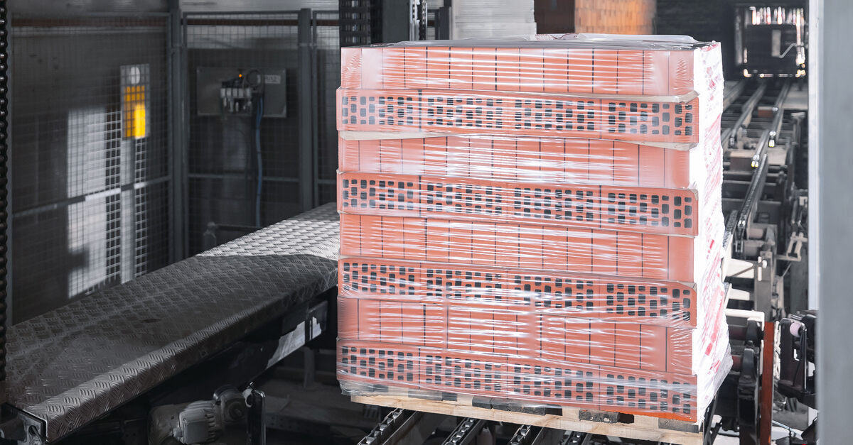 A pallet of red bricks stacked eight bricks high sits on a chain conveyor. The pallet is wrapped in transparent stretch film.