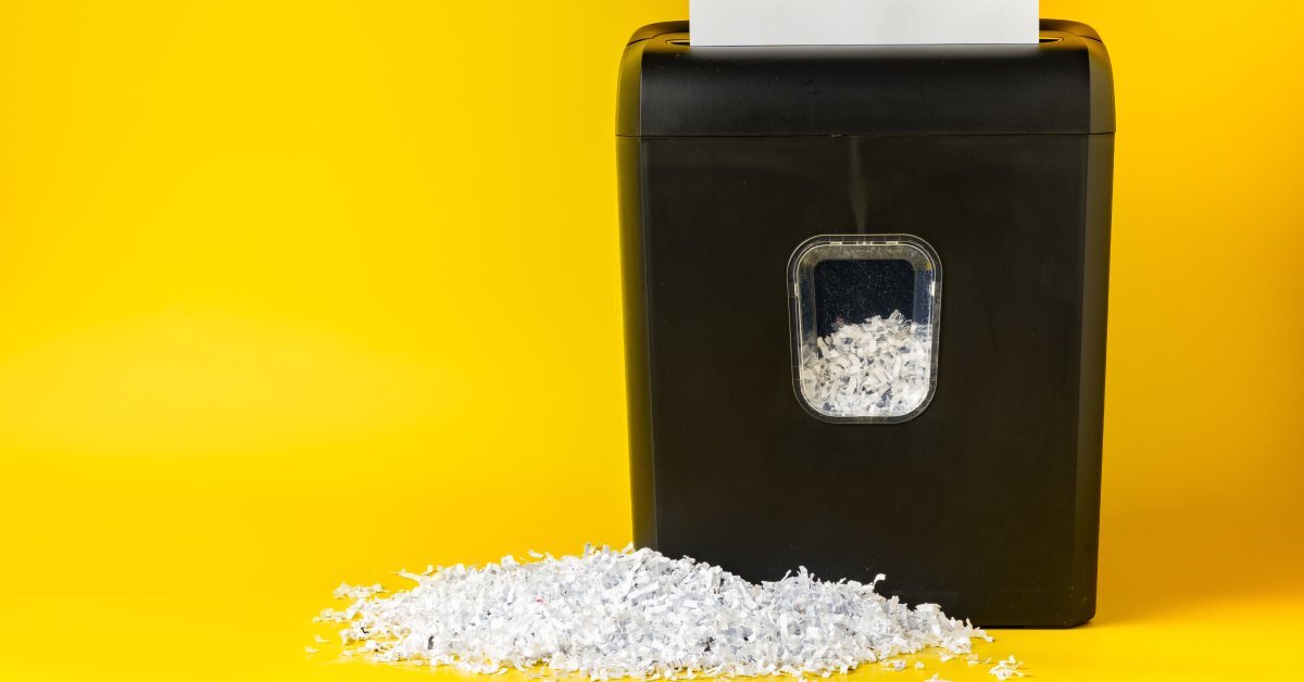 A sheet of paper inserted into the top of a shredder machine. A pile of shredded paper is in front of the machine.