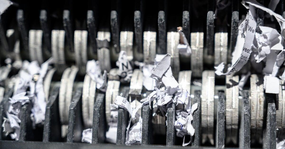 A close-up of the metal teeth on a shredder machine. In the teeth are narrow strips of paper that have partial words on them.