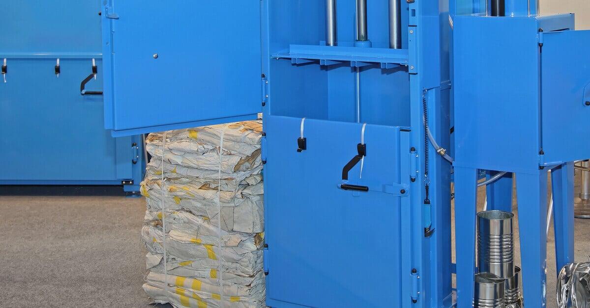 A blue vertical baler with an open door set up near other baling machines. Next to the baler is a tied bale of paper materials.