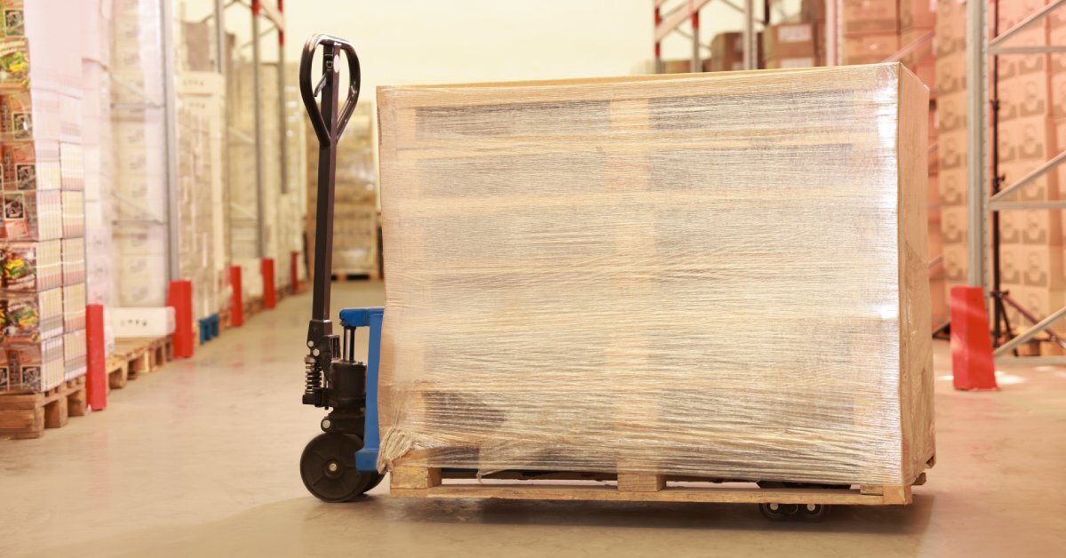 A pallet jack in a warehouse features stacked wooden pallets. The empty pallets are wrapped tightly with stretch wrap.