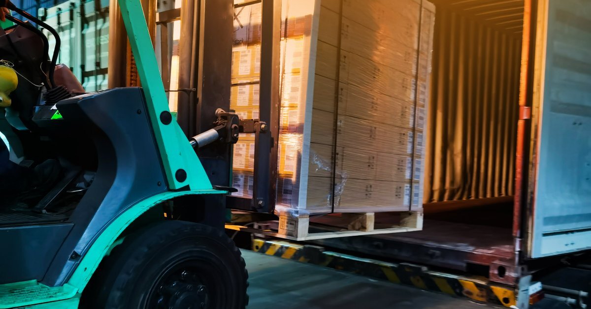 A forklift carries palletized goods into a container. The load is covered with stretch wrap, straps, and corner guards.