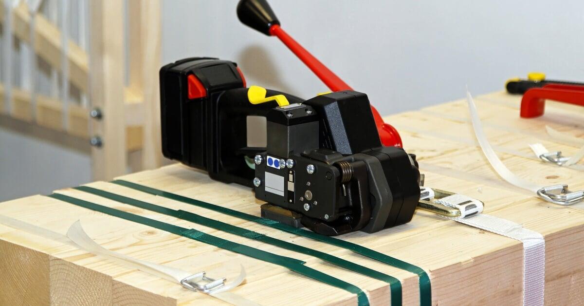 A black and red strapping machine sitting on top of light-colored pieces of wood gathered with white and green straps.