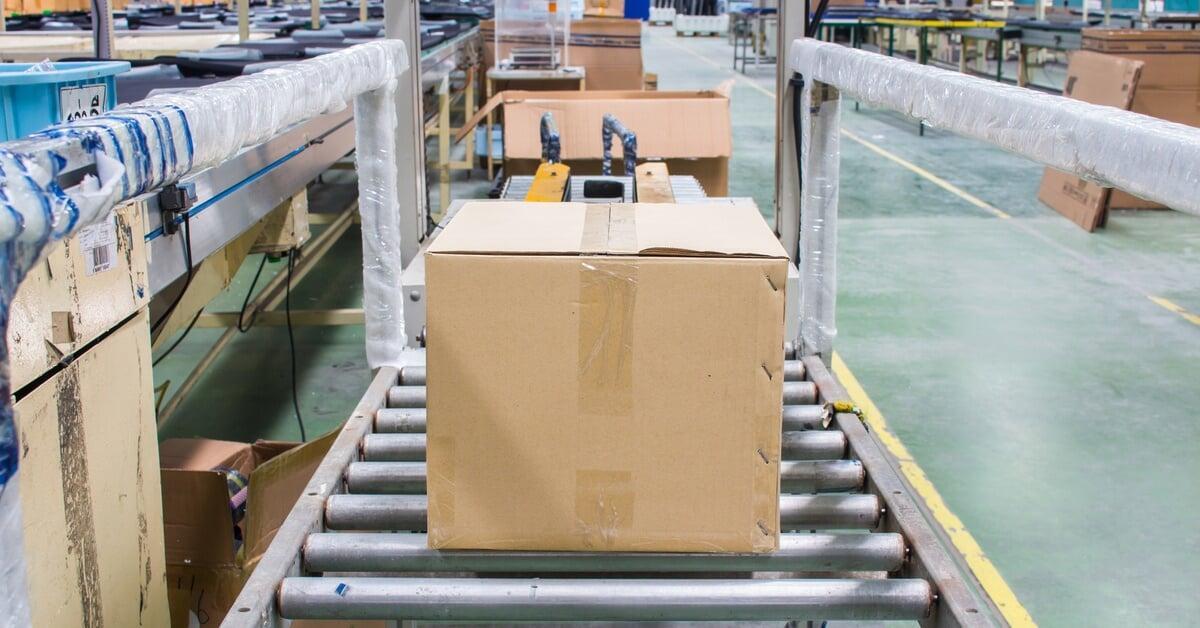 A cardboard box sealed with tape sitting on stainless steel conveyor rollers in a warehouse full of equipment.