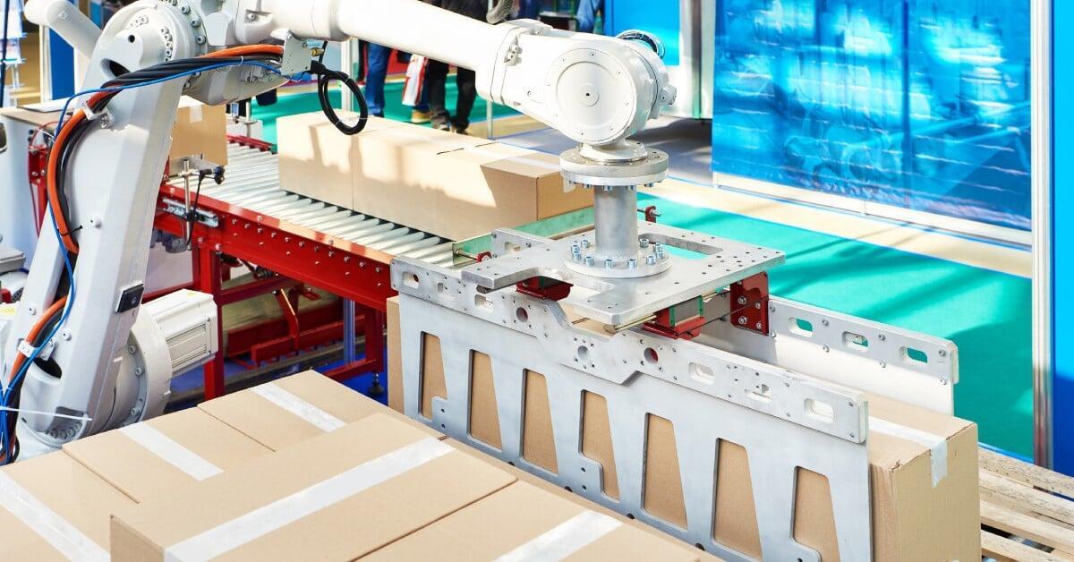 A white robotic arm stacking brown cardboard boxes from a conveyor belt onto a wooden pallet in a warehouse.