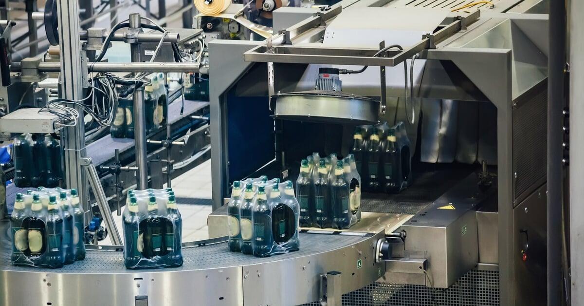 A production line with packs of bottles wrapped in plastic moving through a machine on a conveyor belt.