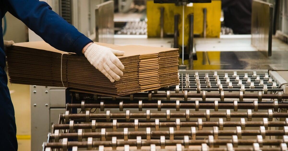 An individual in a blue shirt and white gloves removing a stack of flattened cardboard boxes from a conveyor belt.