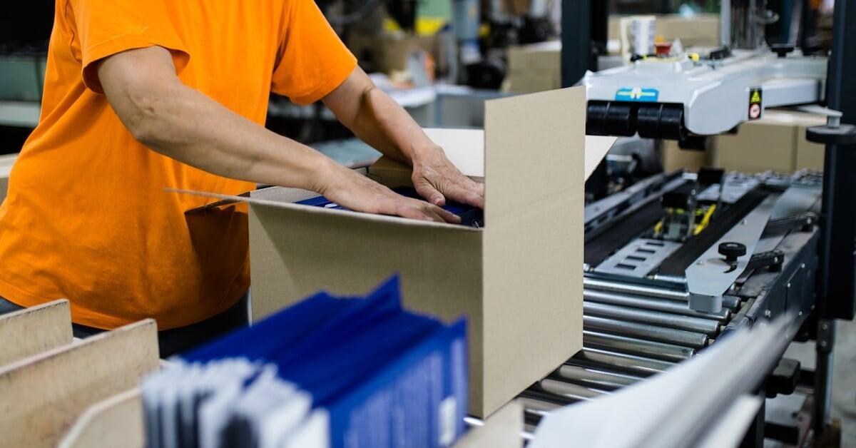 An individual in an orange T-shirt standing in front of a conveyor belt while packing a brown cardboard box.