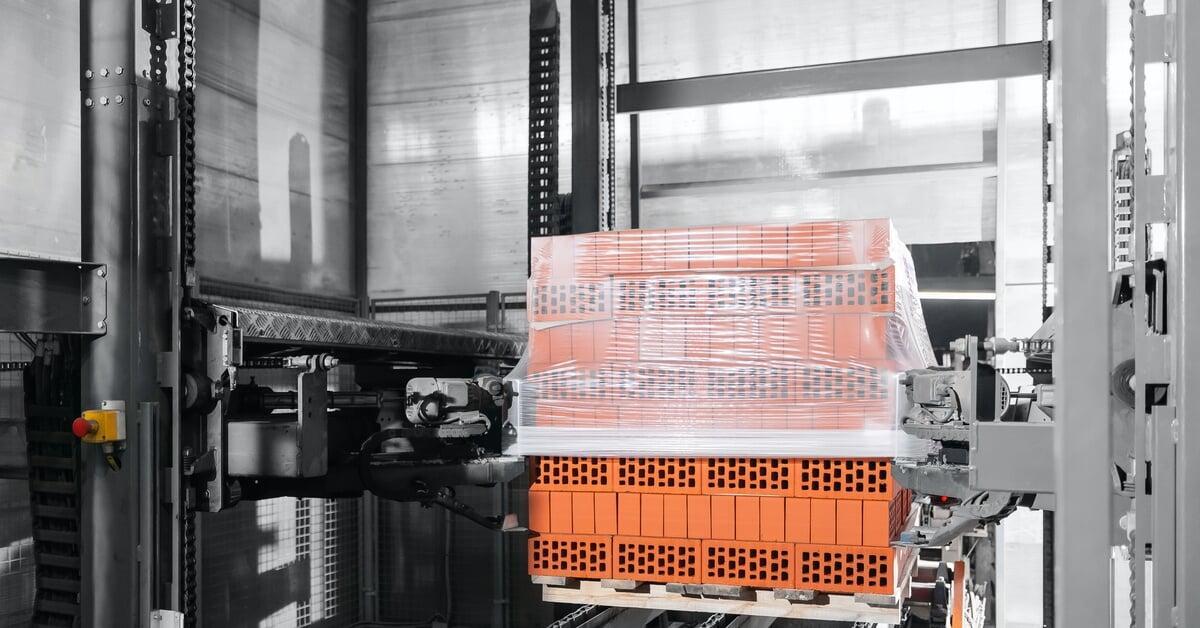 A stretch wrap machine applying stretch film to a stack of orange bricks sitting on a wooden pallet.