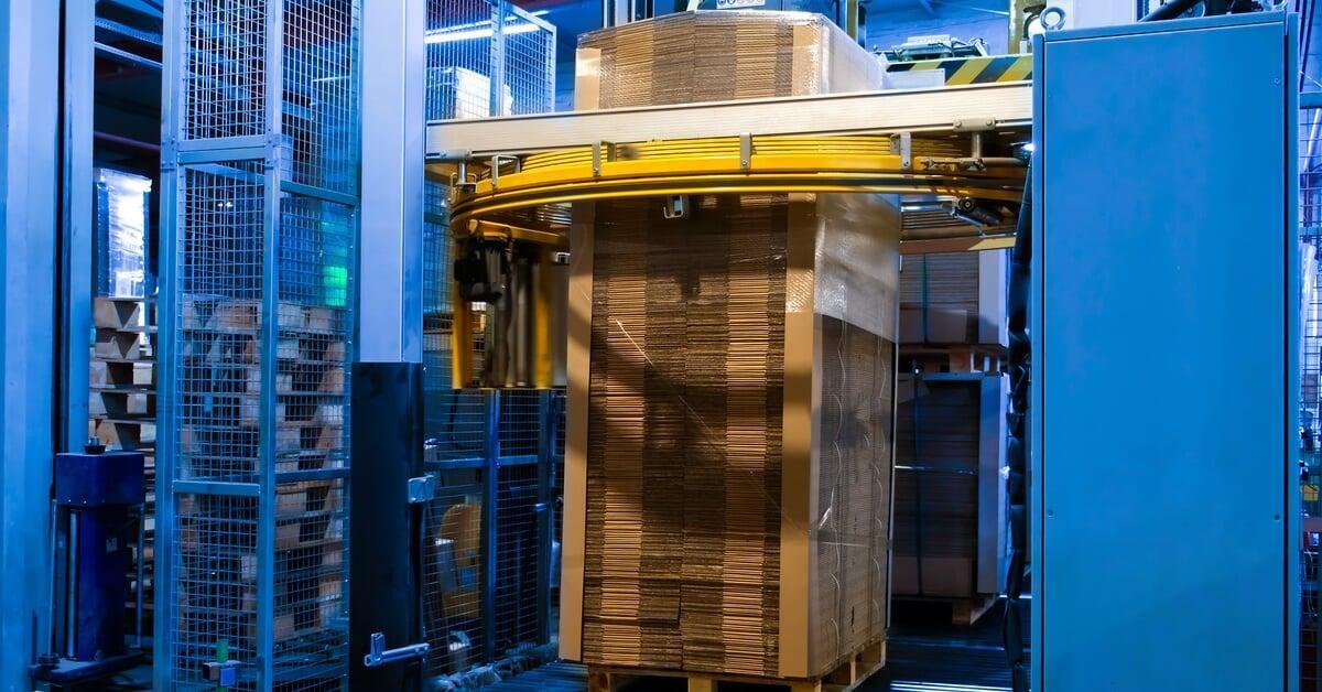 A tall stack of corrugated cardboard sitting on a pallet inside a large blue stretch wrapping machine.