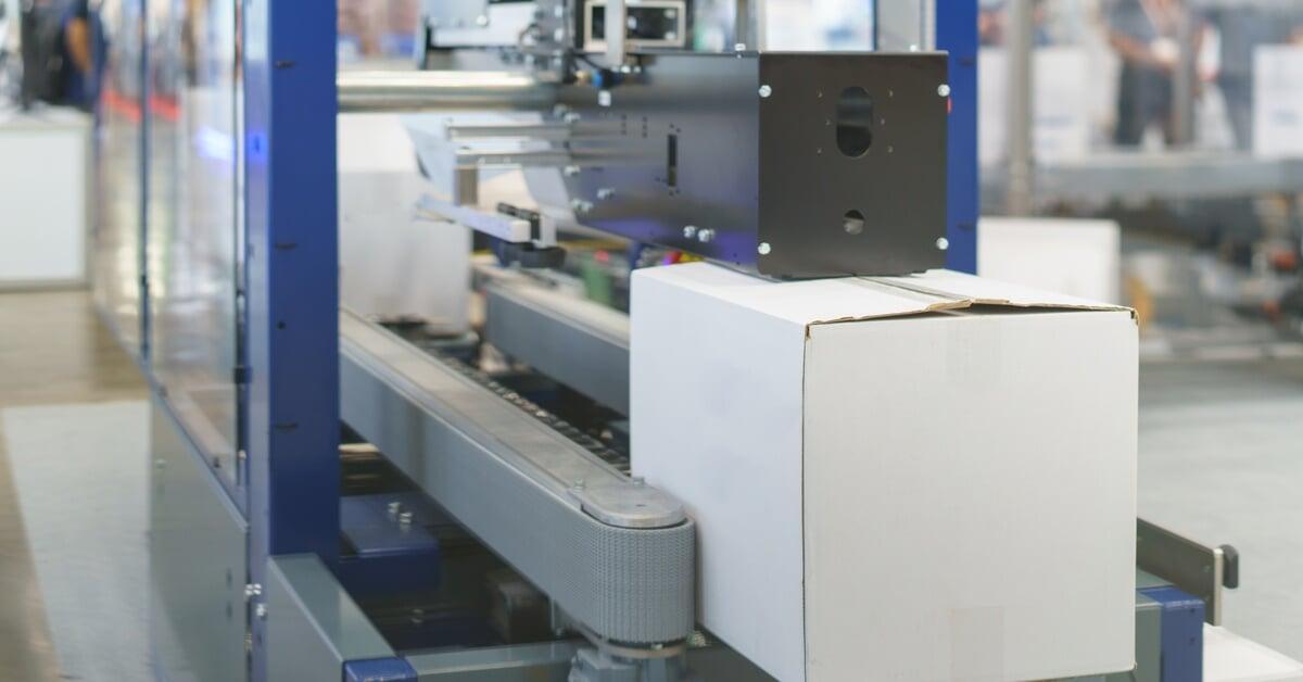 A large blue and gray machine with a roll of tape automatically sealing the top of a white cardboard box.