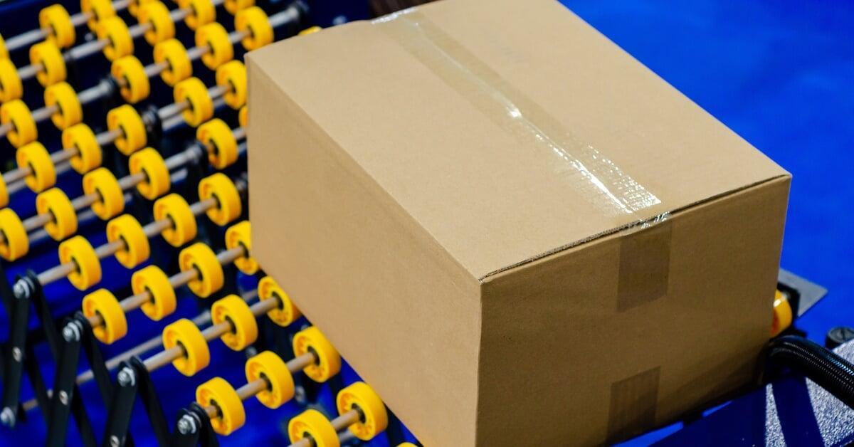 An overhead view of a brown cardboard box sealed with tape sitting on a silver and yellow conveyor belt.
