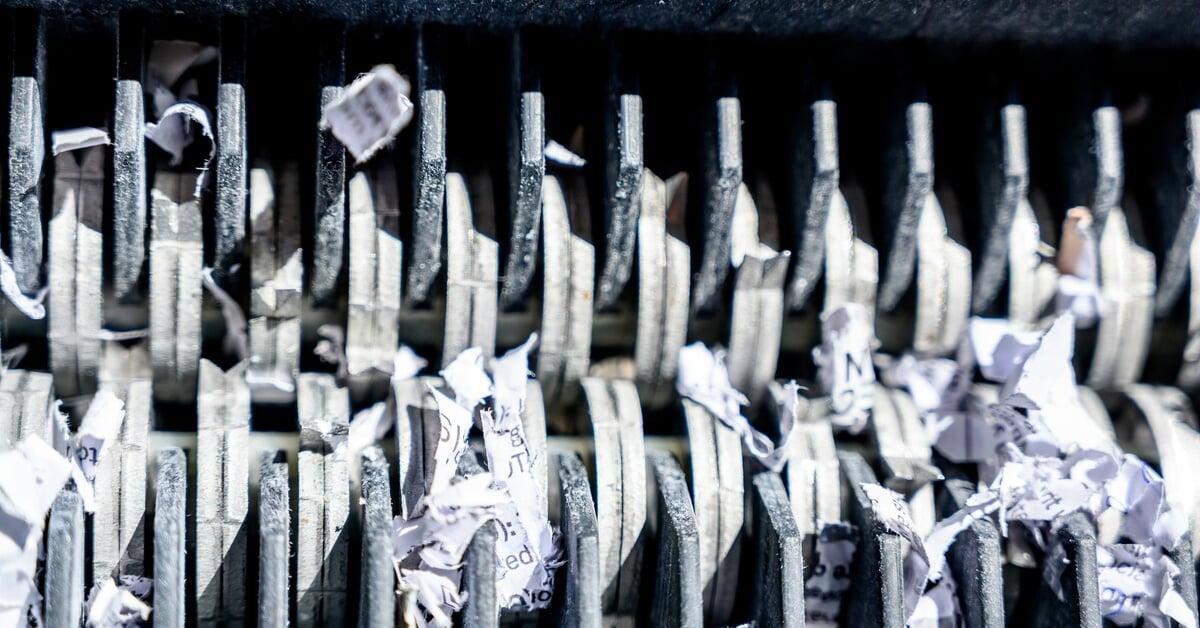 A close-up of the teeth of a paper shredder. There are strips of white paper with black text wedged between the blades.