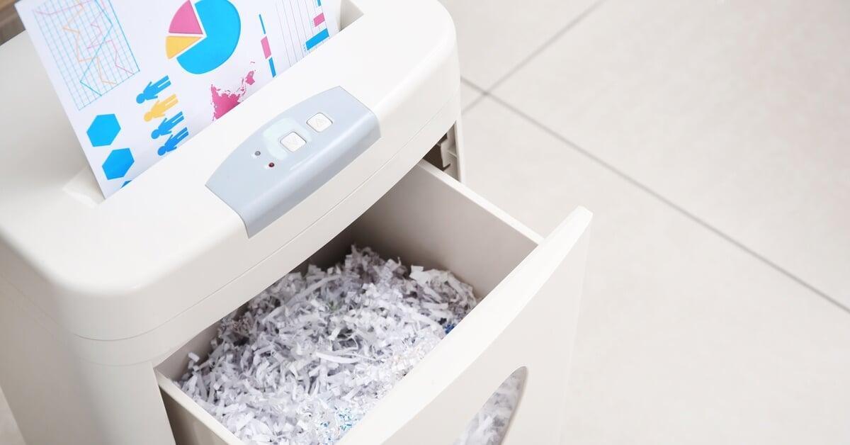 A close-up of a white shredder sitting on a tile floor. There are pieces of shredded paper in the machine's front compartment.