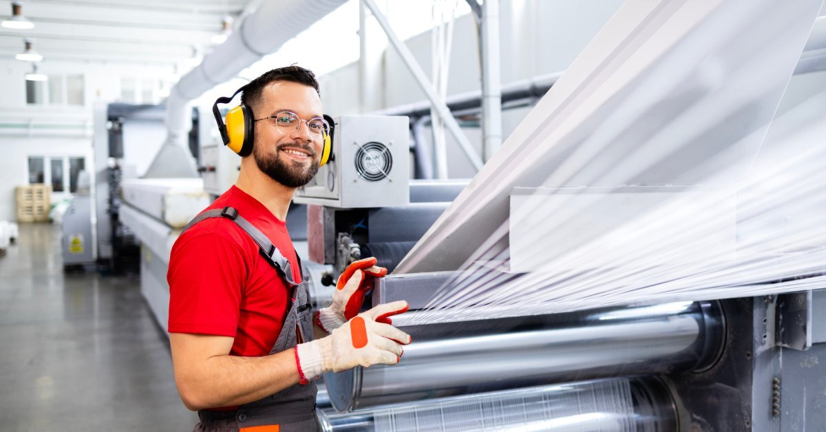 A man in overalls, a red shirt, gloves, and earmuffs standing next to a machine rolling large sheets of plastic.