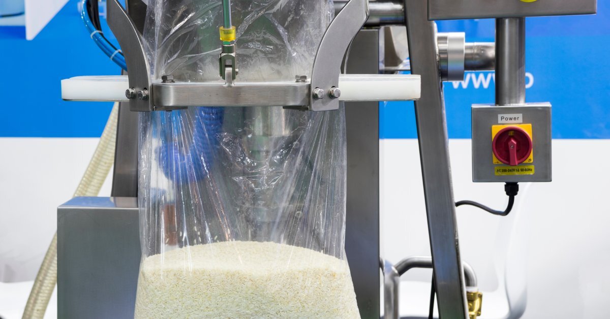 A close-up of a silver packaging machine automatically filling a large plastic bag with rice. The machine has several buttons.