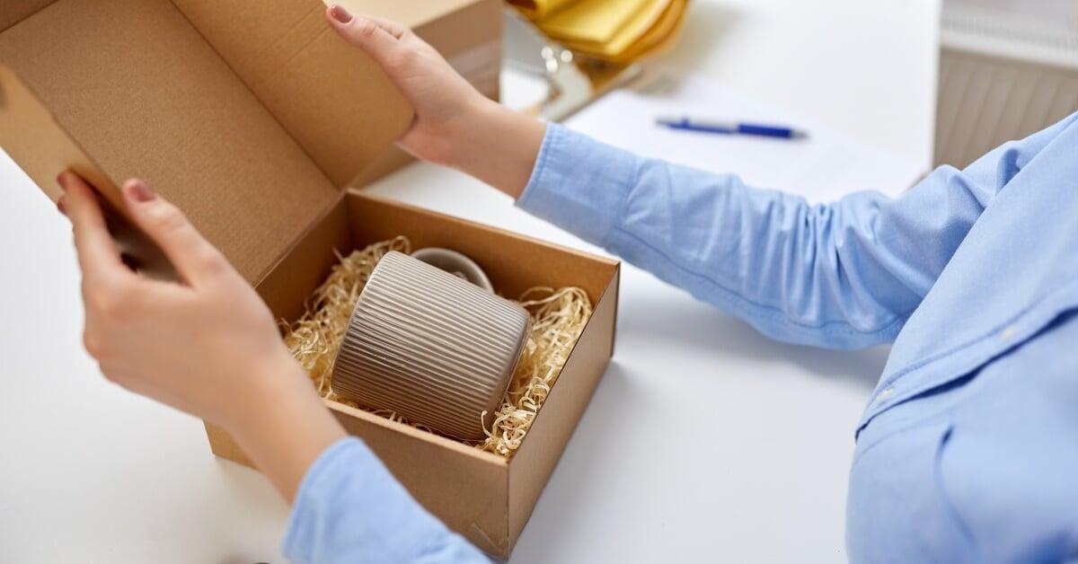 An individual in a blue shirt packaging a brown mug in a cardboard box with paper filler for protection.