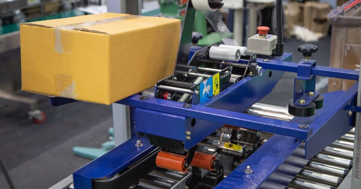 A blue case sealer machine with a sealed cardboard box and a roll of tape sitting on a conveyor belt in a warehouse.