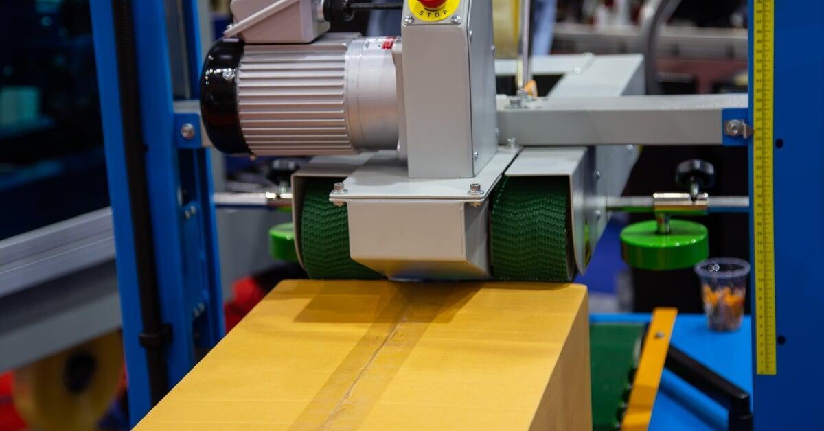 A silver automatic case sealer machine adding a strip of packing tape to the top of a closed cardboard box.