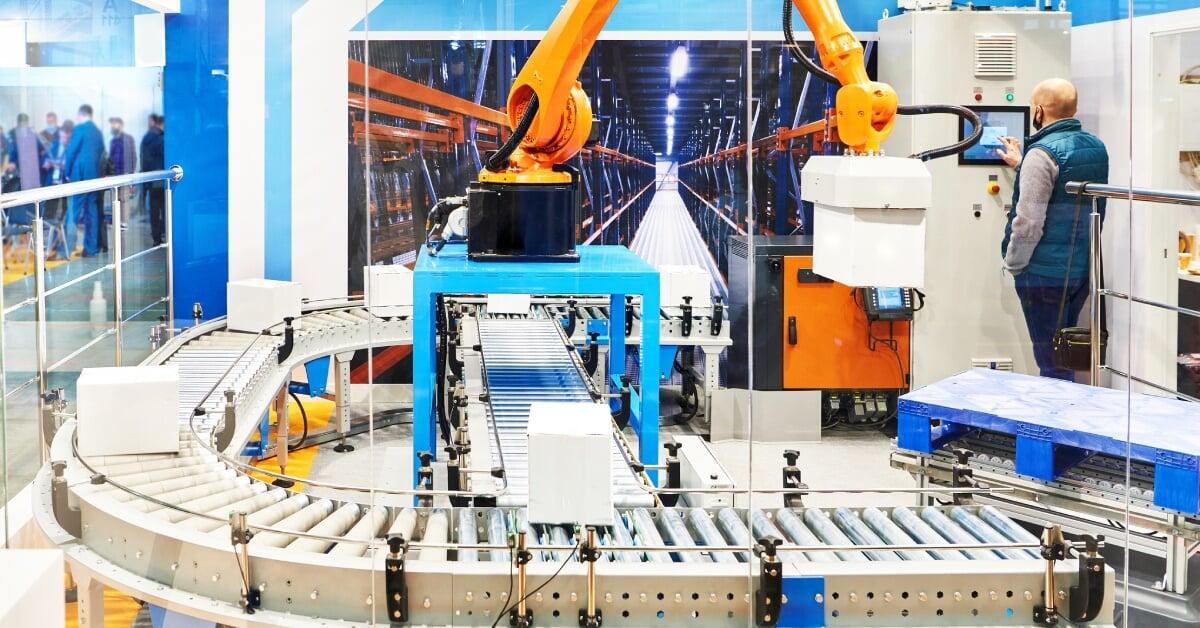 A man using a touchscreen to operate a robotic palletizing machine to lift a white box off a conveyor belt.