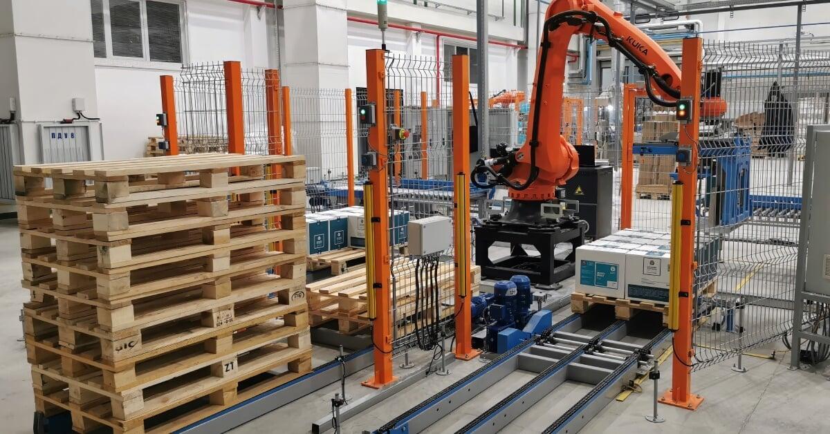 An orange robotic palletizing arm stacking white boxes on a wooden pallet in a warehouse with exposed ducts.