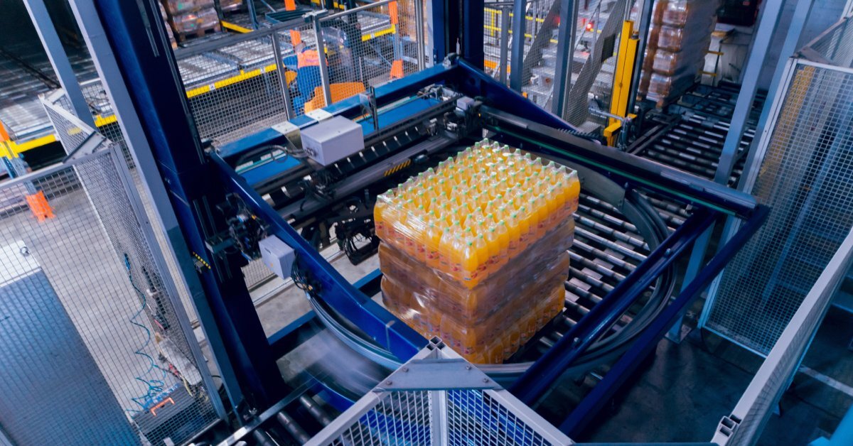 Several layers of orange bottled beverages wrapped in plastic sitting on the conveyor belt of a strapping machine.