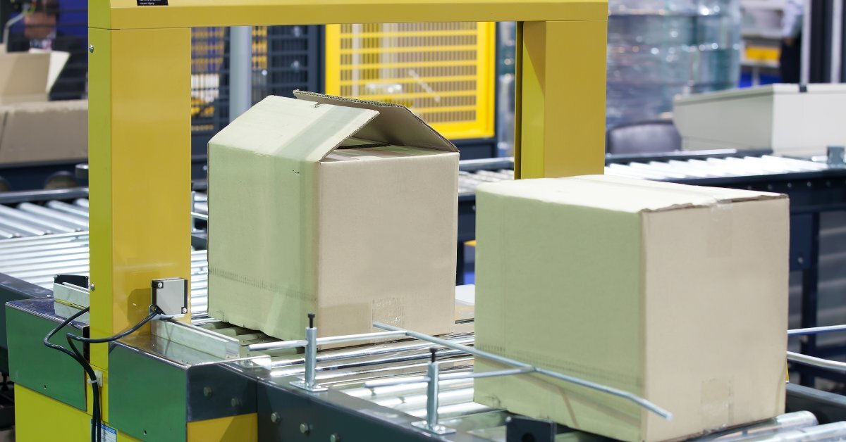 Two cardboard boxes, one open and one closed, sitting on the conveyor belt of a yellow machine in a warehouse.