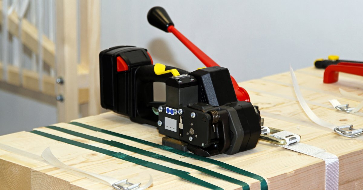 A black and red strapping machine resting on a stack of wooden beams secured with white and green straps.