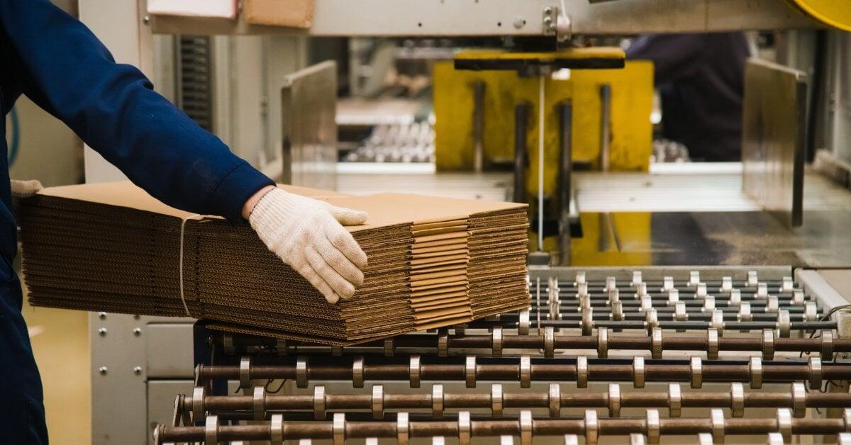  An individual in a blue outfit and white gloves removing a stack of corrugated cardboard from a conveyor belt.