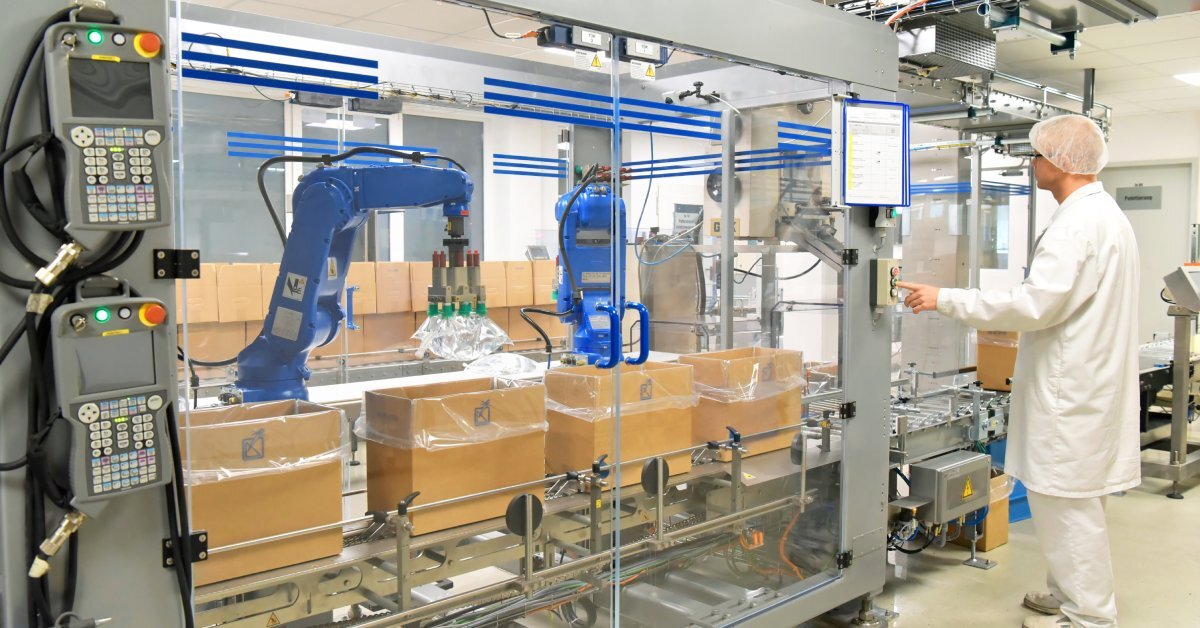 A man in a white uniform and hair net pushing a button to operate packaging equipment with two robotic arms.