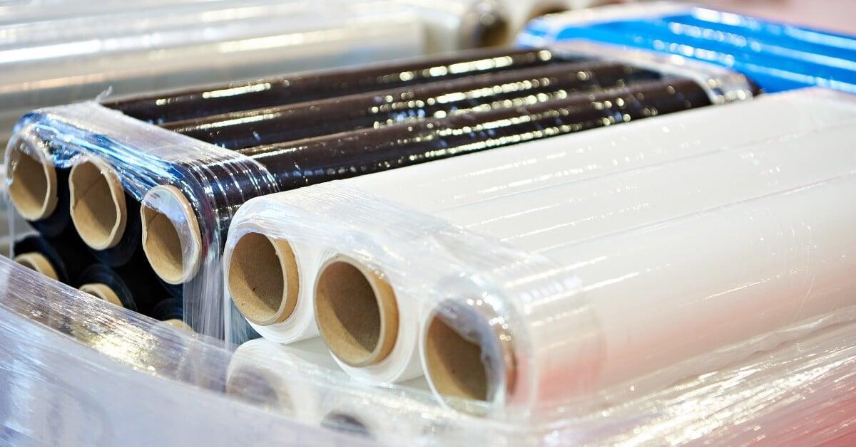 Three black and three clear cellophane wrap rolls stacked on other rolls, with surrounding rolls blurred.