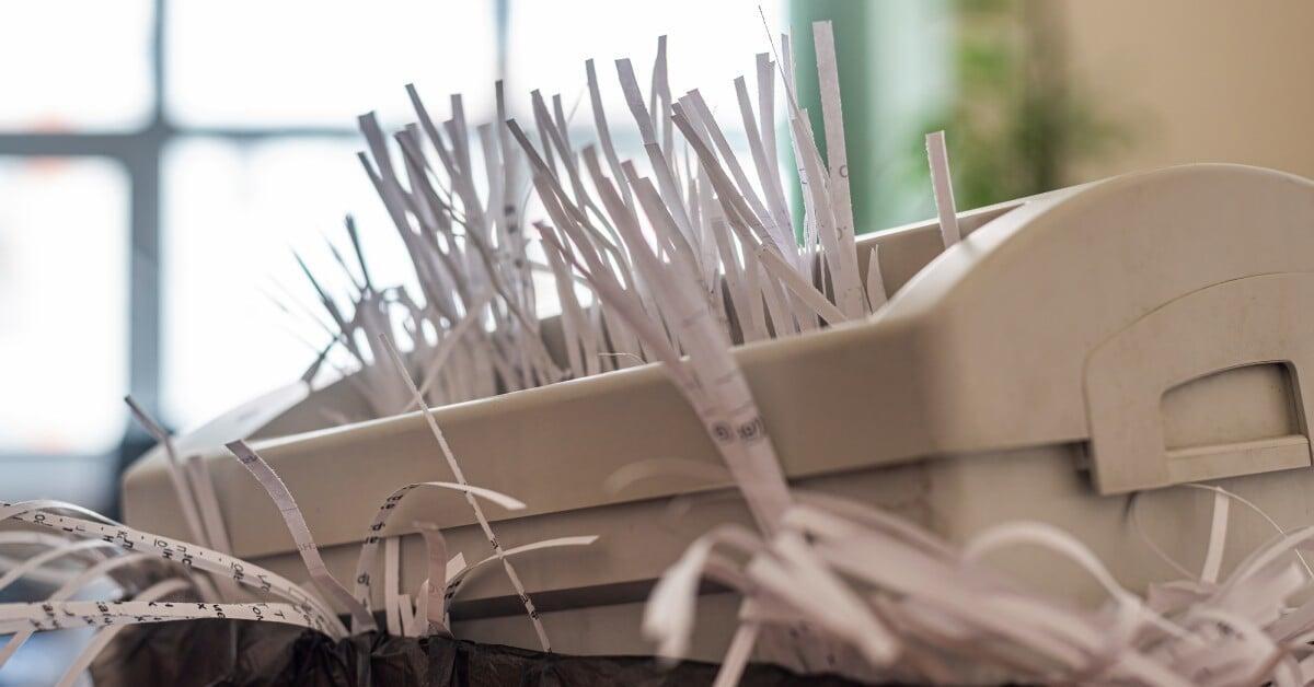A beige office shredder overflowing with shredded paper strips sit above a trash can lined with a black garbage bag.