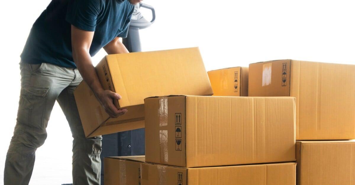 A partially seen worker lifts a packaging box onto pallets in a brightly lit setting. Other boxes also sit on the pallet.